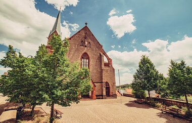 Eine große Kirche aus Buntsandstein mit einem hohen Kirchturm. Ein Platz or der Kirche mit Bäumen. Höpfingen im Odenwald.