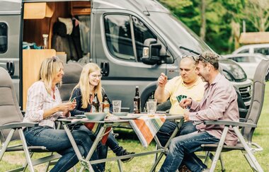 Zwei Frauen und zwei Männer sitzen an einem Campingtisch und spielen Karten. Im Hintergrund ist ein graues Wohnmobil. Odenwald.