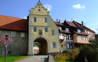 Das Tor ist gelb und hat einen Giebel. Daneben befinden sich Fachwerkhäuser auf der Stadtmauer