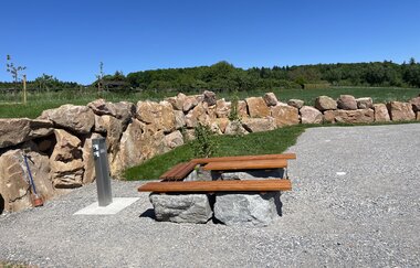 Ein geschotterter Platz mit einer Sitzgelegenheit aus Holz. Die Umgrenzung des Platzes ist aus Findlingsteinen. Im Hintergrund sind Wiesen und Wälder. Limbach im Odenwald.