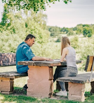 Ein mann und eine Frau sitzen an einem Sitzgelegenheit aus Stein. Im Hintergrund sind Wiesen und Felder. Mudau im Odenwald.