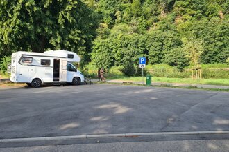 Geteerter Wohnmobilstellplatz mit einem weißen Wohnmobil daneben stehen zwei Frauen. Hinter dem Wohnmobil fliesst der Neckar. | Neckargerach | Odenwald