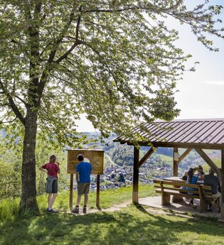 Auf der rechten Seite ist eine Wanderunderstand mit 3 sitzenden Personen. Daneben ist eine Infotafel. Davor stehen ein Mann und eine Frau. Odenwald.