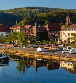 Im Vordergrund ist ein Fluß mit einem Binnenschiff. Dahinter am Ufer ist direkt ein Wohnmobilstellplatz mit 3 Wohnmobilen. Im Hintergrund ist eine Stadt mit Häusern und einer Kirche. Eberbach im Odenwald.