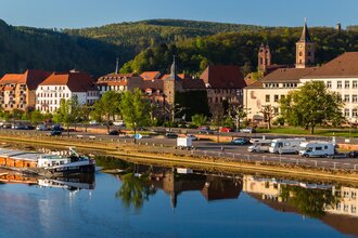 Im Vordergrund ist ein Fluß mit einem Binnenschiff. Dahinter am Ufer ist direkt ein Wohnmobilstellplatz mit 3 Wohnmobilen. Im Hintergrund ist eine Stadt mit Häusern und einer Kirche. Eberbach im Odenwald.