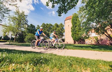 Zwei Radfahrer auf einem Radweg. Im Hintergrund ist eine Stadtmauer mit einem Turm. Eberbach im Odenwald.