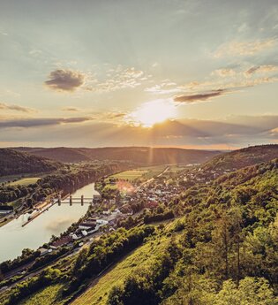 Weitblick mit Sonnenuntergang auf einen Fluss. Auf der rechten Uferseite erstreckt sich eine Gemeinde. Neckarzimmern im Odenwald.