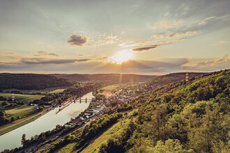 Weitblick mit Sonnenuntergang auf einen Fluss. Auf der rechten Uferseite erstreckt sich eine Gemeinde. Neckarzimmern im Odenwald.
