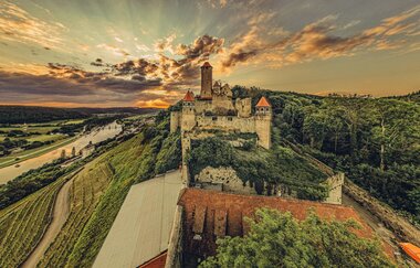 Blick auf eine Burg die auf einem Berg steht. Auf der linken Seite fließt ein Fluss auf der rechten Seite ist ein Wald. Burg Hornberg im Odenwald.