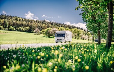 Weißes Wohnmobil fährt auf einer Strasse. Im Vordergrund ist eine Blumenwiese im Hintergrund eine Steuobstwiese mit angrenzendem Wald. | Odenwald