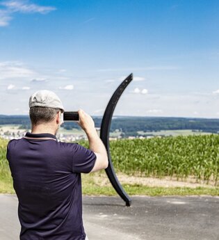 Ein Mann mit Fernglas schaut in die Ferne. Vor ihm stehen zwei halbrunde Gebilde, die aussiehen wie eine große Klammer. Dahinter ist ein grünes Feld. Osterburken im Odenwald.