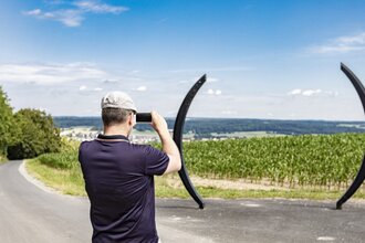 Ein Mann mit Fernglas schaut in die Ferne. Vor ihm stehen zwei halbrunde Gebilde, die aussiehen wie eine große Klammer. Dahinter ist ein grünes Feld. Osterburken im Odenwald.
