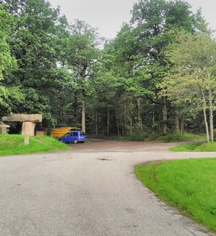 Ein Parkplatz am Waldrand. Auf dem Platz stehen zwei Fahrzeuge. Davor führt ein Weg vorbei. Schönbrunn im Odenwald.