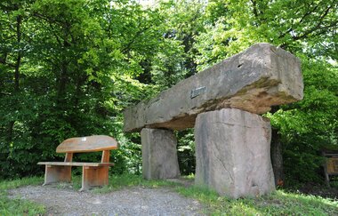 Man sieht einen kleinen Platz im Wald mit einer Holzbank. Daneben stehen 3 große Findlinge die wie eine überdimensionale Brücke angeordnet sind. Schönbrunn im Odenwald