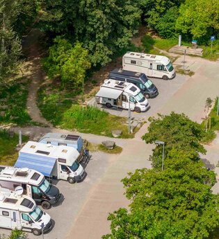 Wohnmobilstellplatz mit mehreren geparkten Wohnmobilen, umgeben von Bäumen.