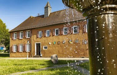 Man sieht ein Schloss mit kleinem Türmchen und Holzläden an den Fenstern. Im Vordergrund sieht man ein Teil eines Brunnen mit perlendem Wasser. Odenwald.