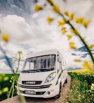 Ein weisses Wohnmobil auf einer Straße. Links ist ein grünes Feld auf der rechten Seite ein gelbes Rapsfeld. Odenwald.