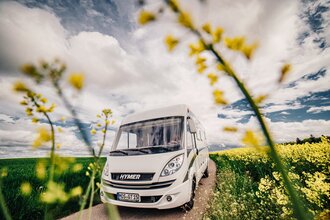 Ein weisses Wohnmobil auf einer Straße. Links ist ein grünes Feld auf der rechten Seite ein gelbes Rapsfeld. Odenwald.