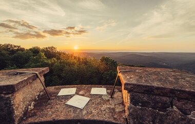 Man sieht den Rand eines Steinturmes mit 3 Plaketten und Blick in die Weite und den Sonnenuntergang. Odenwald.