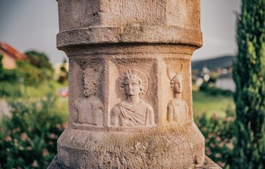 Ein Ausschnitt einer Buntsandsteinsäule mit römischen Männern. Im Hintergrund eine verschwommene Stadt mit Wäldern. Odenwald.