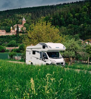 Im Vordergrund ist eine grüne Wiese mit einem Wohnmobil zu sehen. Im Hintergrund ein Schloss. Es handelt sich um das Schloss Zwingenberg am Neckar im Odenwald.