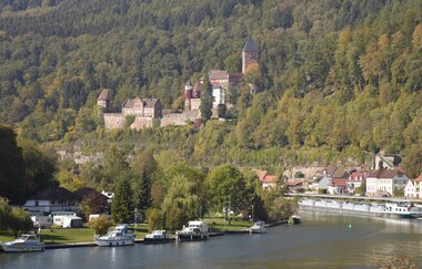 Blick auf eine Flußbierung mit Booten am linken Ufer. Im Hintergrund ist ein großen Schloß auf einem bewaldeten Hügel zu sehen. Zwingenberg im Odenwald.