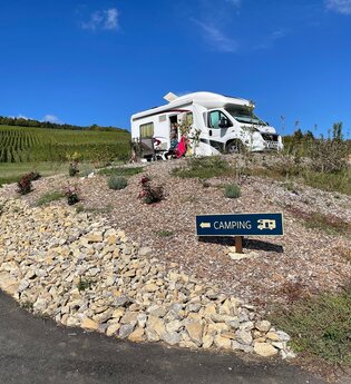 Blick auf den am Rand der Weinberge gelegenen Wohnmobilstellplatz Weingut Ehrmann.  Hier steht ein Wohnmobil. Bei schönstem Wetter und blauem Himmel genießen die im Freien sitzenden Gäste den Ausblick.