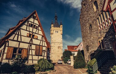 Blick in eine historische Gasse in der Altstadt Bad Wimpfen. Im Vordergrund ein Fachwerkhaus und ein Steingebäude, im Hintergrund ein historischer Burgturm aus Stein.