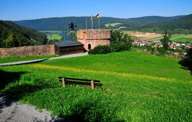 Blick auf den Turm mit Hexe bei der Freudenburg. Im Hintergrund ist Freudenberg, im Vordergrund eine Bank mit Wiesezu sehen.