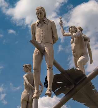 Am Ortseingang von Lauffen am Neckar erhebt sich das eindrucksvolle Kunstwerk „Hölderlin im Kreisverkehr“ des Bildhauers Peter Lenk. Auf einem geschwungenen „H“ balanciert eine überdimensionale Schreibfeder, auf der Friedrich Hölderlin als Kind und als ju