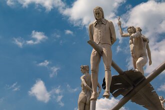 Am Ortseingang von Lauffen am Neckar erhebt sich das eindrucksvolle Kunstwerk „Hölderlin im Kreisverkehr“ des Bildhauers Peter Lenk. Auf einem geschwungenen „H“ balanciert eine überdimensionale Schreibfeder, auf der Friedrich Hölderlin als Kind und als ju