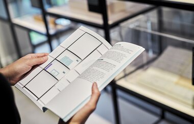 Eine Person hält eine Broschüre in den Händen, dahinter sind Regale mit Ausstellungsstücken