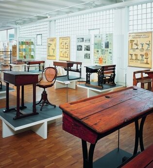 Blick in das Schulmuseum der Firma VS in Tauberbischofsheim. Hier sind alte Schulbänke, Tische und Stühle sowie Wandkarten zu sehen.