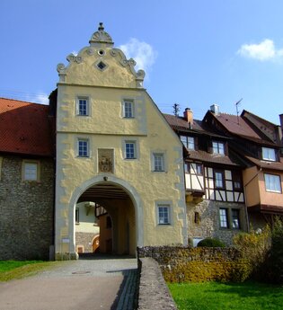 Das Tor ist gelb und hat einen Giebel. Daneben befinden sich Fachwerkhäuser auf der Stadtmauer
