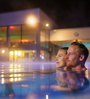 Ein Man und eine Frau sind im Mineralaußenbecken der Therme bei Nacht.