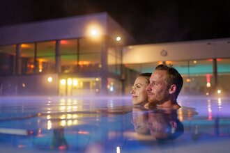 Ein Man und eine Frau sind im Mineralaußenbecken der Therme bei Nacht.