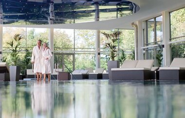 Im Innenraum der Therme spiegelt das Wasser die Fenster. Hinten steht ein Pärchen in Bademanteln.
