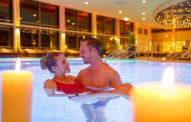 Ein Mann und eine Frau befinden sich im Wasser, der Mann hält die Frau in seinen Armen. Der Innenraum der Therme erstrahlt im Kerchenlicht.