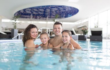 Eine Familie mit zwei jungen Mädchen schaut in die Kamera und lächelt. Sie stehen im Wasser der Innenbecken.