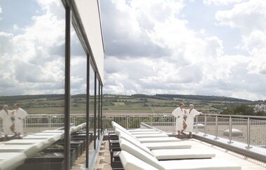 Auf der Dachterrasse befindet sich der Ruhebereich der Saunalandschaft. Bequeme Liegen laden zum Ausruhe ein, eine Frau und eine Mann stehen weiter hinten in Bademänteln.