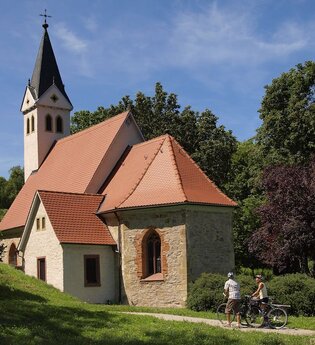 Zwei Radfahrer stehen vor einer Kapelle mit einem spitzen Turm
