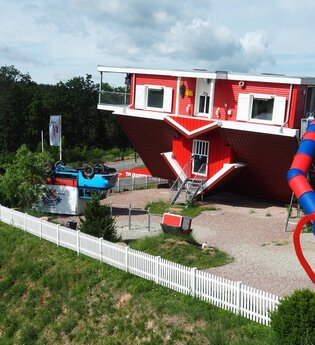 Blick auf das Toppels-Haus "Verdrehte Welt". Das Haus und ein Auto davor stehen auf  dem Kopf. Im Außenbereich ist ein Spielplatz mit Spielgeräten, z.T. auch auf dem Kopf stehend, zu sehen.
