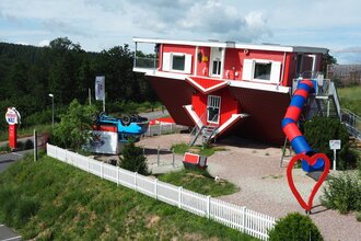 Blick auf das Toppels-Haus "Verdrehte Welt". Das Haus und ein Auto davor stehen auf  dem Kopf. Im Außenbereich ist ein Spielplatz mit Spielgeräten, z.T. auch auf dem Kopf stehend, zu sehen.