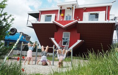 Blick auf Spielgeräte vor dem Toppels-Haus "Verdrehte Welt". Hier hängen drei Kinder mit dem Kopf nach unten an Kletterstangen.