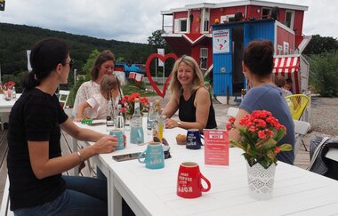 Blick auf mehrere Personen, die vor dem Toppels-Haus "Verdrehte Welt" Getränke zu sich nehmen. Die Tassen stehen ebenfalls auf dem Kopf.