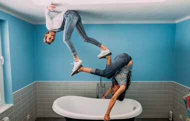 Blick auf zwei Frauen über einer Badewanne. Die eine hängt vermeintlich mit dem Rücken an der Decke, die andere hält sich mit den Händen an der Badewanne fest und der Rest des Körpers zeigt Richtung Decke. Die Füße der beiden berühren sich.