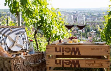 Mittig ist eine umgedrehte Weinkiste. Darauf stehen zwei Weingläser mit Rotwein und ein Teller mit Brot,  Käse und Trauben. Daneben steht ein geöffneter Korb, in dem Geschirr zu sehen ist. Im Hintergrund sieht man Weinreben und eine Stadt von oben.