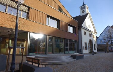 Außenansicht der Tourist-Info mit Glasfront und moderner Holzfassade. Vor den Gebäude stehen Bänke zum Verweilen.