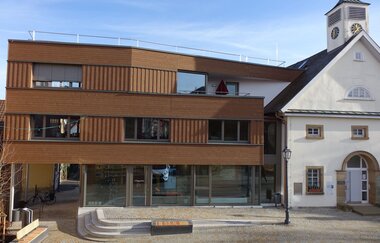 Außenansicht der Tourist-Information über den Obertorplatz. Das Gebäude hat eine moderne Holzfassade sowie eine große Glasfront. Rechts neben der Tourist-Info ist das Theodor-Heuss-Museum zu sehen.