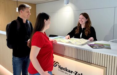 Beratung eines Paares an der Theke der Tourist-Info. Vor den Gästen und der Mitarbeiterin liegt eine Karte und diverse Prospekte.